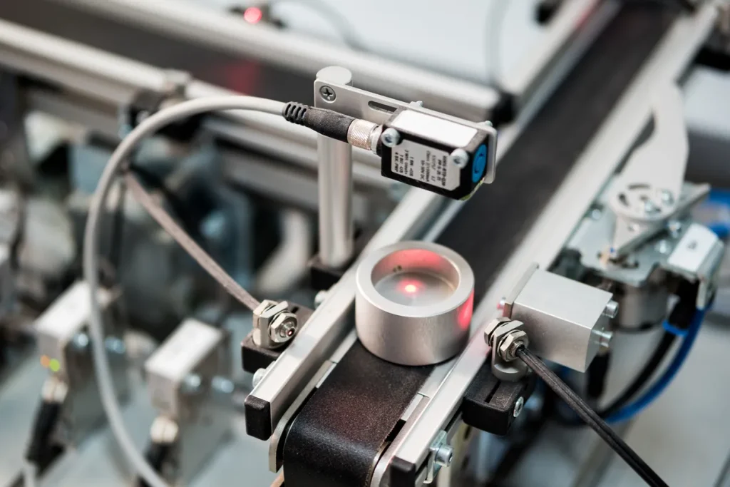 Inline optical sensor illuminating a metal part on a conveyor, the mounting arrangement that shows how a color sensor works in production.