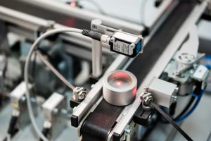 Inline optical sensor illuminating a metal part on a conveyor, the mounting arrangement that shows how a color sensor works in production.